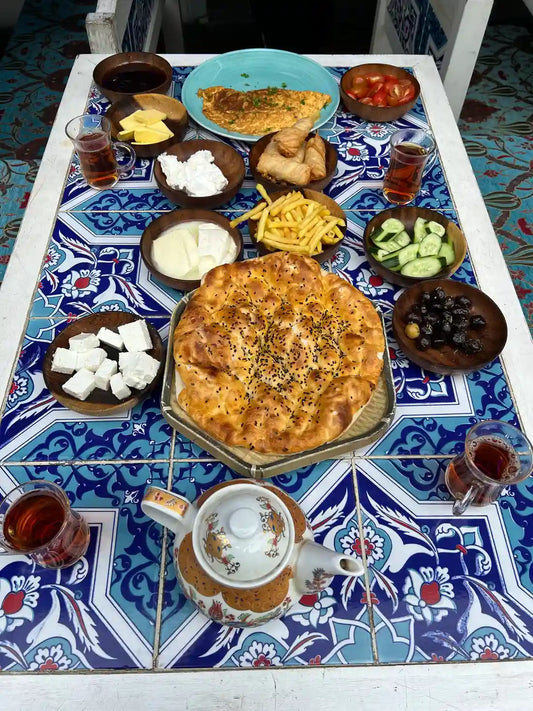 A lavish Traditional Turkish Breakfast spread (Serpme Kahvaltı) centered around a fresh sesame flatbread, surrounded by assorted cheeses, olives, pastries, and tulip-shaped tea glasses on a blue-tiled table.